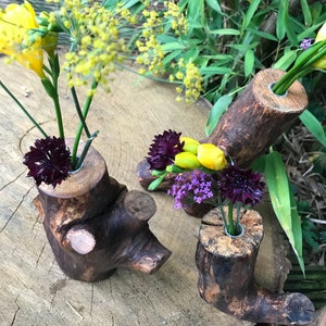 May include: Three rustic wooden log vases with different sizes and shapes, each with a hole drilled in the top for flowers. The vases are filled with yellow, purple, and dark purple flowers.