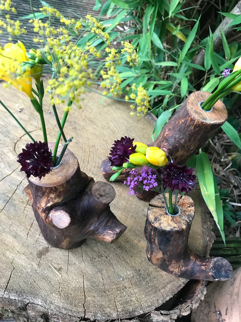 May include: Three rustic wooden log vases with different sizes and shapes, each with a hole drilled in the top for flowers. The vases are filled with yellow, purple, and dark purple flowers.