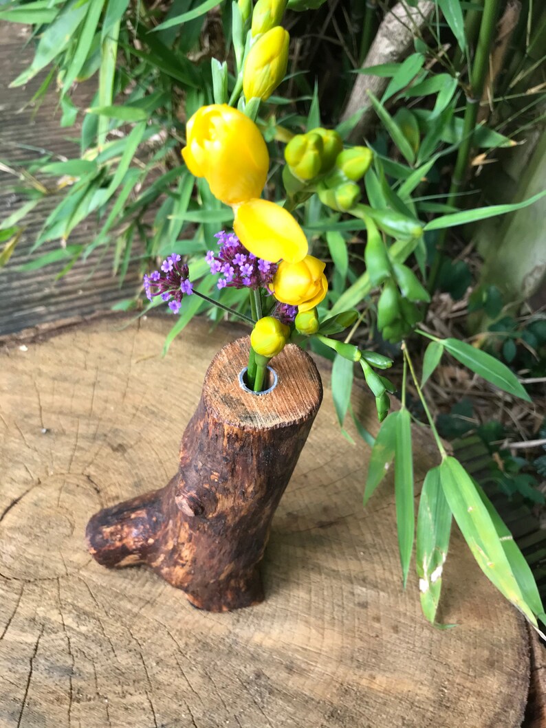 May include: A rustic wooden vase with a natural bark finish, featuring a single hole at the top for holding flowers. The vase is filled with yellow and purple flowers.