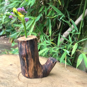 May include: A small wooden vase carved from a tree trunk with a natural brown and black finish. The vase is shaped like a boot and has a small opening at the top. A single stem of purple flowers is in the vase.