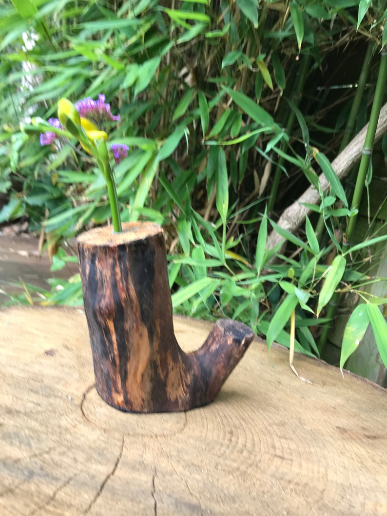May include: A small wooden vase carved from a tree trunk with a natural brown and black finish. The vase is shaped like a boot and has a small opening at the top. A single stem of purple flowers is in the vase.