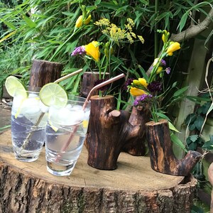May include: Three rustic wooden vases with a natural bark finish, each holding a bouquet of yellow and purple flowers. The vases are arranged on a wooden slice, with two clear glass cups filled with a sparkling beverage and lemon slices.