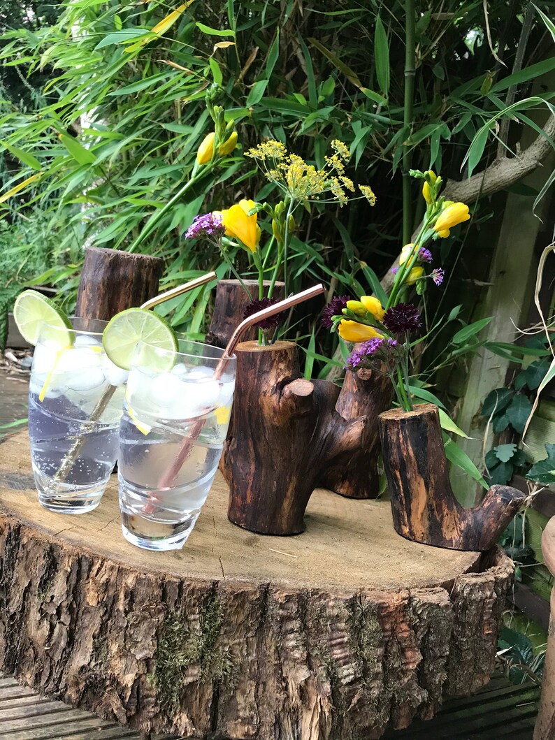 May include: Three rustic wooden vases with a natural bark finish, each holding a bouquet of yellow and purple flowers. The vases are arranged on a wooden slice, with two clear glass cups filled with a sparkling beverage and lemon slices.