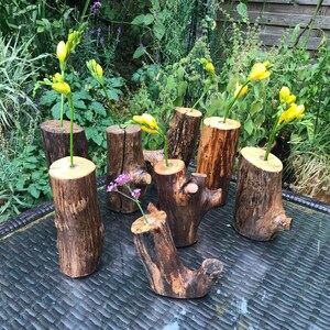 May include: Six rustic wooden vases, each with a different shape and size, are arranged on a black wicker table. The vases are filled with yellow and purple flowers.
