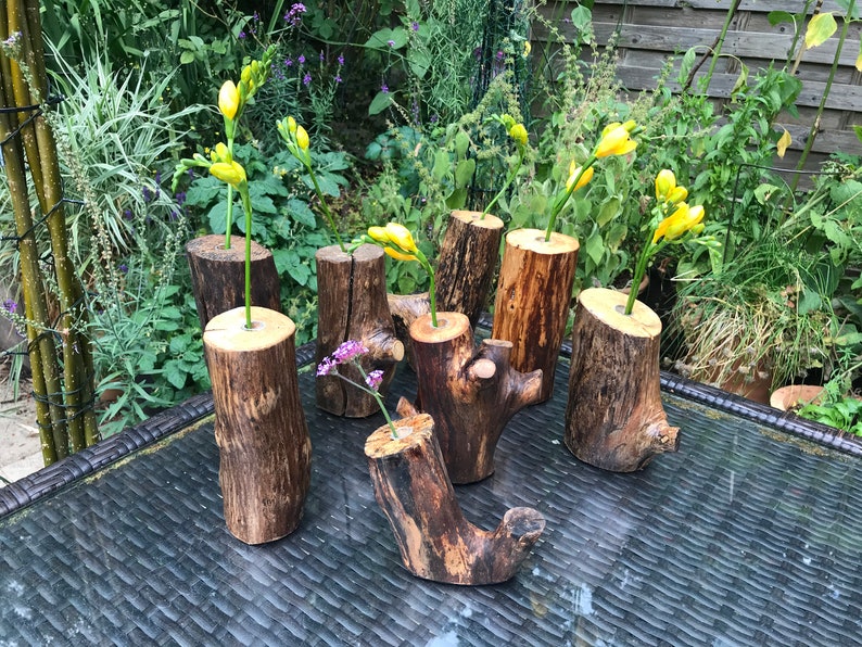 May include: Six rustic wooden vases, each with a different shape and size, are arranged on a black wicker table. The vases are filled with yellow and purple flowers.