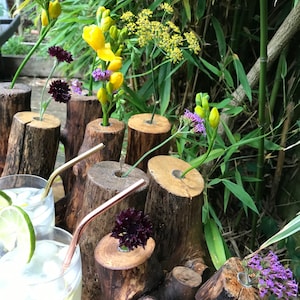 May include: A rustic wooden log vase with multiple openings for flowers. The vase is filled with yellow freesia, purple flowers, and green foliage. The vase is sitting on a wooden surface.