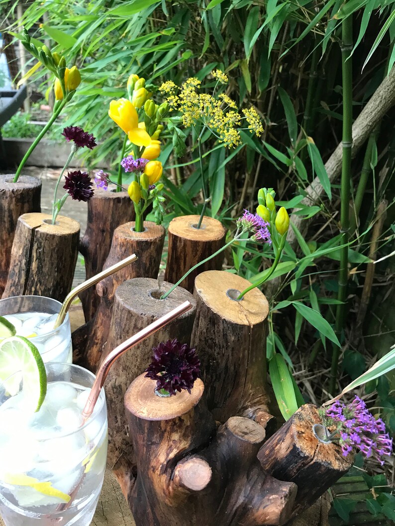 May include: A rustic wooden log vase with multiple openings for flowers. The vase is filled with yellow freesia, purple flowers, and green foliage. The vase is sitting on a wooden surface.