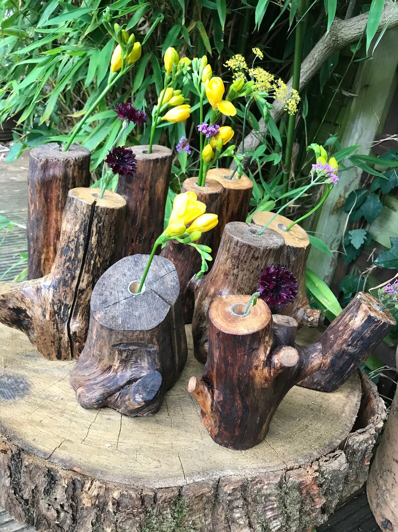 May include: A collection of rustic wooden vases, each with a unique shape and texture, are arranged on a wooden slab. The vases are filled with yellow and purple flowers.