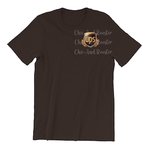 Op de afbeelding: Een bruin t-shirt met een goud en bruin UPS-logo op de borst. Het logo heeft de vorm van een schild en heeft een decoratieve rand. De tekst "Chic And Rooster" is op het shirt gedrukt.