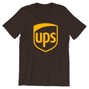Peut inclure: Un t-shirt marron foncé avec le logo UPS. Le logo est un bouclier doré avec les lettres "ups" en or. Le t-shirt est fait d'une matière douce et a des manches courtes.
