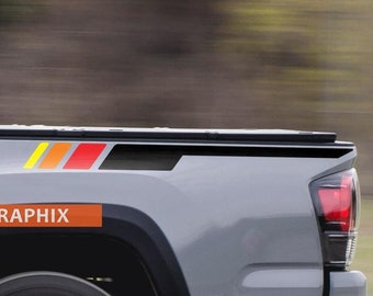 May include: A grey pickup truck with black and red racing stripes on the side of the bed. The truck is driving on a tarmac road. The text "SUPDEC" and "GRAPHIX" is visible on the truck.