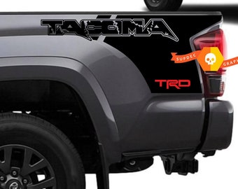 May include: Black and grey truck bed side decals with the word "TACOMA" in white lettering and the word "TRD" in red lettering. The decals are designed to be applied to the side of a truck bed.