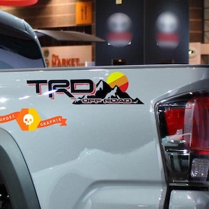 May include: A grey pickup truck with a black "TRD OFF ROAD" decal on the side. The decal features a mountain range with a yellow and orange sunset. The truck also has a black and orange skull graphic decal with the text "SUPDEC GRAPHIX".