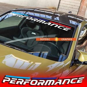 May include: A black car windscreen with a white "PERFORMANCE" decal featuring blue, red and white stripes. The decal is positioned at the top of the windscreen.