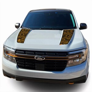 May include: A white Ford pickup truck with a black grille and a gold patterned hood decal. The decal has a swirling design and is on both sides of the hood.