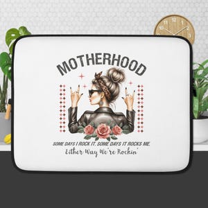 May include: White laptop sleeve with a black border featuring a graphic of a woman with sunglasses and a leather jacket. The text "MOTHERHOOD" is above the image, with the quote "SOME DAYS I ROCK IT. SOME DAYS IT ROCKS ME. Either Way We're Rockin'" below.