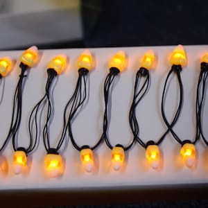 May include: A string of candy corn-shaped lights with black wires. Each light has a yellow and white color scheme, with a bright orange glow emanating from within. The lights are arranged in a row on a white surface, perfect for Halloween decorations.