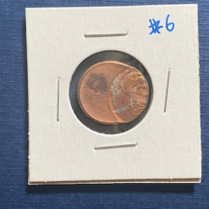 May include: A copper-colored US penny with the words "In God We Trust" and "Liberty" visible. The coin is mounted on a white card with the number "#6" written in blue ink.