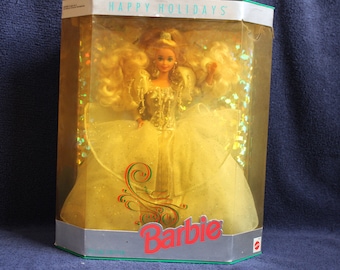 May include: A Barbie doll in a white gown with gold accents, wearing a tiara and holding a bouquet of flowers. The doll is in a clear plastic box with a gold and green background. The box says "Happy Holidays" and "Barbie" on it.