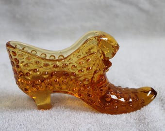 May include: Amber-colored glass shoe figurine with a textured, hobnail design. The decorative shoe has a pointed toe, a small heel, and a curved top. A charming collectible item.