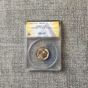 May include: A 1938-D/S 5C Buffalo nickel, graded MS 65, encased in a clear protective holder. The coin features a profile of a Native American on one side. The holder includes the text "1938-D/S 5C BUFFALO FS-512 OMM-2" and "MS 65".