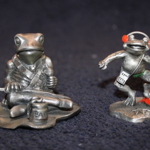 May include: Two pewter frog figurines. One frog is seated, holding a drumstick and a small barrel that says "BA". The other frog is dancing, wearing headphones and a cassette player. The dancing frog is standing on a red surface.