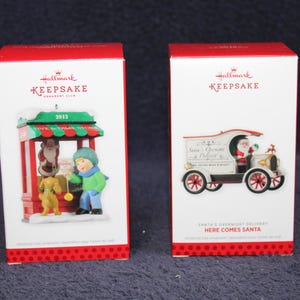 May include: Two Hallmark Keepsake Christmas ornament boxes. One features a boy, dog, and shop scene, the other a vintage Santa delivery truck. Both boxes are white with red accents and the Hallmark Keepsake logo.