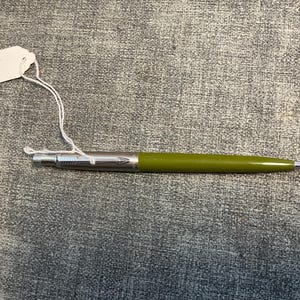 May include: A green and silver pen with a white tag attached. The pen has a silver tip and a green barrel. The tag is blank. The pen is lying on a gray textured surface.