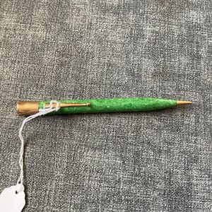 May include: A vintage pen with a green speckled barrel and gold-colored accents. The pen has a gold-colored tip and a gold-colored clip. A white tag is attached to the pen with a white string.