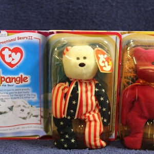 May include: Two Ty Beanie Babies in their original packaging. The first is a white bear wearing a red, white, and blue outfit with stars and stripes. The second is a red bear with a gold ribbon around its neck. The packaging includes the text "International Bears II" and "Spangle the Bear".