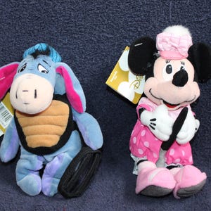 以下が含まれることがあります： ディズニーキャラクターのぬいぐるみ2体。黒とオレンジのストライプのシャツを着た青とピンクのイーヨーのぬいぐるみ。白のドット柄のピンクのドレスとピンクのリボンをつけたピンクと黒のミニーマウスのぬいぐるみ。