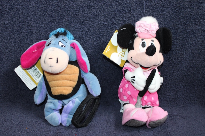 以下が含まれることがあります： ディズニーキャラクターのぬいぐるみ2体。黒とオレンジのストライプのシャツを着た青とピンクのイーヨーのぬいぐるみ。白のドット柄のピンクのドレスとピンクのリボンをつけたピンクと黒のミニーマウスのぬいぐるみ。