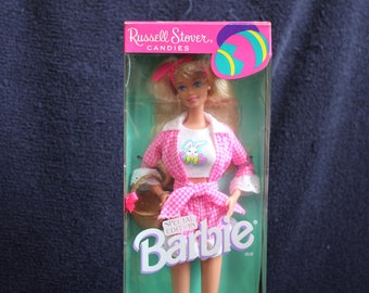 May include: A pink and white gingham doll with blonde hair and a pink headband. The doll is holding a basket and is wearing a white shirt with a bunny on it. The doll is in a box with the words "Russell Stover Candies" and "Barbie" on it.