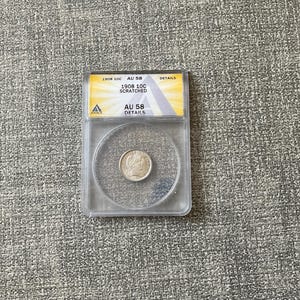 May include: A silver coin encased in a clear plastic holder. The coin is centered within the holder. The holder has a yellow and white label with the text "1908 10C SCRATCHED AU 58 DETAILS". The coin is graded AU 58.