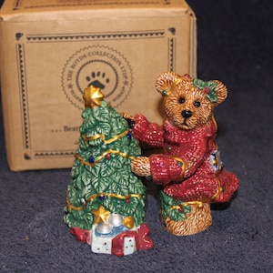 May include: A red and gold Christmas bear figurine holding a green Christmas tree figurine decorated with gold stars and ornaments. The figurines are part of The Boyds Collection Ltd.
