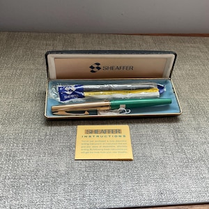 May include: A vintage Sheaffer pen set in a black box with a gold trim. The set includes a green and gold pen, ink cartridges, and a small instruction card. The box is open, revealing the contents on a blue interior.