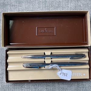 May include: A vintage Sheaffer pen set in a brown box. The set includes a silver-colored pen and a silver-colored mechanical pencil. The box has a brown lid with the Sheaffer logo and a beige interior. A tag with the number 752 is attached.