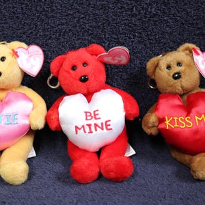 May include: Three plush teddy bears with heart-shaped patches. The bears are brown, red, and tan. The hearts have the words "Cutie", "Be Mine", and "Kiss Me" on them.