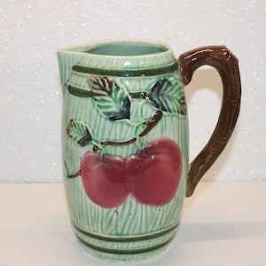 May include: A green ceramic pitcher with a brown handle. The pitcher is decorated with a vine of green leaves and two red apples.