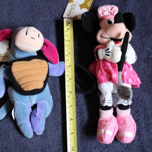 以下が含まれることがあります： ディズニーキャラクターのぬいぐるみ2体。ピンクの尻尾と背中に茶色の甲羅が付いた青いイーヨー。白地にピンクのドット柄のピンクのドレスを着て、黒いゴルフクラブを持ったピンクと白のミニーマウス。