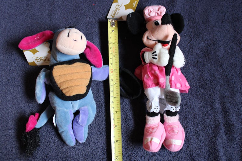 以下が含まれることがあります： ディズニーキャラクターのぬいぐるみ2体。ピンクの尻尾と背中に茶色の甲羅が付いた青いイーヨー。白地にピンクのドット柄のピンクのドレスを着て、黒いゴルフクラブを持ったピンクと白のミニーマウス。