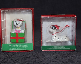 May include: Two Dalmatian dog Christmas ornaments in their original packaging. One ornament features a Dalmatian dog wearing a Santa hat and peeking out of a green gift box with a red ribbon. The other ornament features a Dalmatian dog wearing a red Santa hat and lying down. Both ornaments have a red string attached.