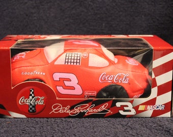 May include: A red, white, and black plush toy car with the number 3 on the side. The car is a replica of Dale Earnhardt's NASCAR race car, sponsored by Coca-Cola. The car is in a clear plastic box with a red, white, and black checkered pattern on the bottom.