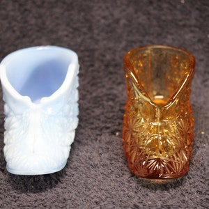 May include: Two glass slipper-shaped trinket dishes. One is a light blue milk glass with a floral pattern. The other is a clear amber glass with a starburst pattern.