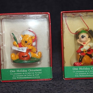 May include: Two boxed Christmas ornaments, one featuring Winnie the Pooh holding a jar of honey and the other featuring Mickey Mouse dressed as a Christmas elf. Both ornaments are made of painted resin and are packaged in clear plastic boxes with red and white labels that read "One Holiday Ornament".