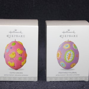 May include: Two Hallmark Keepsake Easter egg ornaments. The ornament on the left is pink with yellow chicks and colorful polka dots. The ornament on the right is purple with a floral design. Both ornaments have the text "Hallmark Keepsake" on the box.