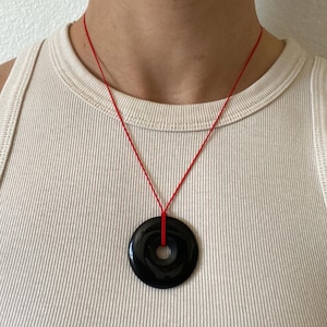 May include: A black circular pendant necklace with a hole in the center, suspended from a thin red cord. The pendant is glossy and reflects light. The necklace is worn against a cream-colored tank top.