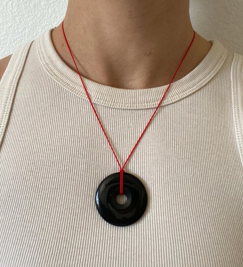 May include: A black circular pendant necklace with a hole in the center, suspended from a thin red cord. The pendant is glossy and reflects light. The necklace is worn against a cream-colored tank top.