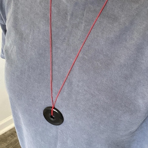 May include: A black circular pendant necklace with a red cord. The pendant has a hole in the center. The necklace is hanging against a blue t-shirt.