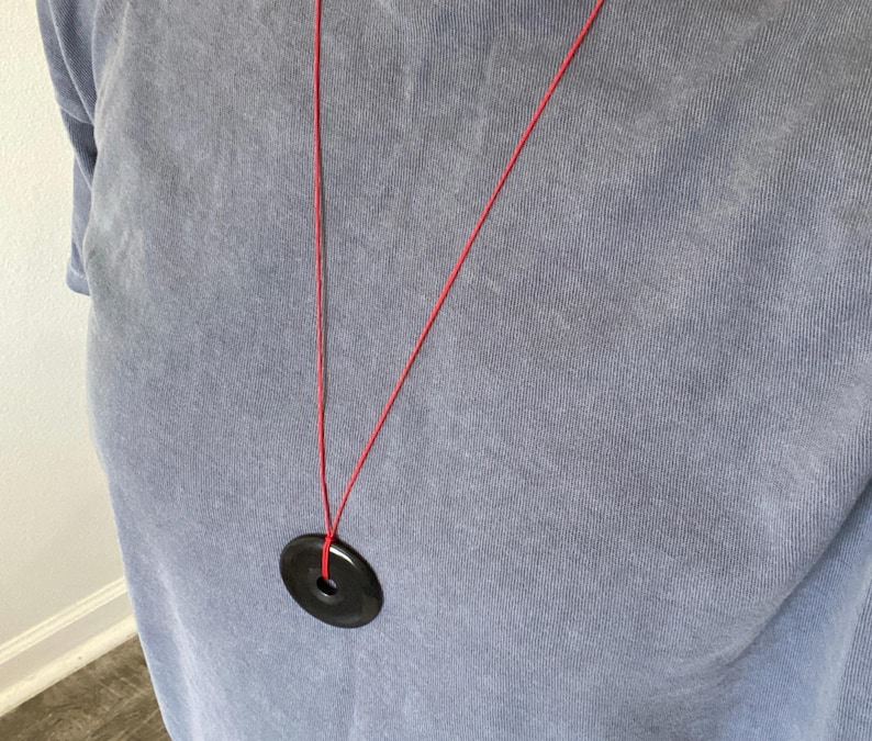 May include: A black circular pendant necklace with a red cord. The pendant has a hole in the center. The necklace is hanging against a blue t-shirt.